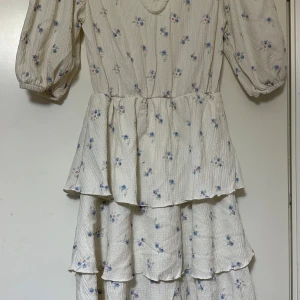 Vit blommig klänning med volanger - Charmig vit klänning med små blommor i blått.. Klänningen har trekvartsärmar och flera lager av volanger som ger en romantisk känsla. Perfekt för vår och sommar!