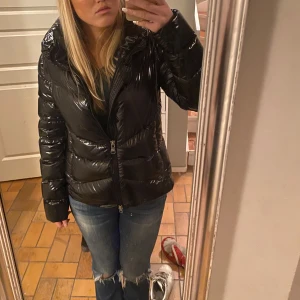 Svart glansig pufferjacka COLMAR - Snygg svart glansig pufferjacka med dragkedja. Perfekt för kyligare dagar med sin vadderade design. Jackan har en normal passform och långa ärmar, jätte snygg