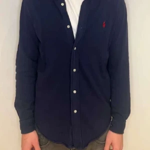 Mörkblå skjorta från Ralph Lauren - Snygg mörkblå skjorta från Ralph Lauren med klassisk krage och knappar framtill. Skjortan har en broderad logga på bröstet och långa ärmar. Perfekt för en stilren look.