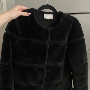 Svart pälsjacka från Neo Noir - Snygg svart pälsjacka från Neo Noir. Jackan har en stilren design med långa ärmar och en mjuk, fluffig yta. Perfekt för att hålla sig varm och trendig under kyliga dagar.