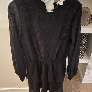 Svart klänning med volangdetaljer - Snygg svart klänning med diskret prickigt mönster och volangdetaljer framtill. Klänningen har långa ärmar och en bekväm passform som passar perfekt för olika tillfällen.