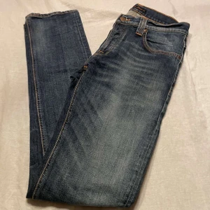 Blå jeans från Nudie Jeans - Säljer ett par snygga blå jeans från Nudie Jeans. De har en klassisk femficksdesign med orange sömmar och en läderpatch med logga på baksidan. Tillverkade i ekologisk bomull för en skön och hållbar känsla. Jeansen är straight!