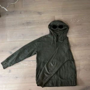 Grön huvtröja med dragkedja - Säljer en grön cp company tröja med dragkedja och inbyggda glasögon. Köpte den här på plick men är lite för stor. Tröjan har långa ärmar och ribbade muddar. Perfekt för en unik stil!
