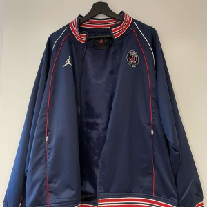 Blå jacka från Jordan x PSG - Säljer en snygg blå jacka från Jordan i samarbete med PSG. Jackan har vita och röda detaljer samt texten 'PARIS' på ryggen. Den har en dragkedja framtill och ribbade muddar vid kragen och ärmsluten. 