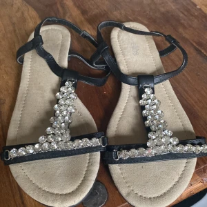 Svarta sandaler med glittriga detaljer - Snygga svarta sandaler med glittriga stenar på remmarna. Perfekta för att ge en touch av glamour till din outfit. Sandalerna har en bekväm sula och justerbara remmar för bästa passform.