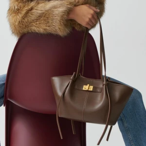 Brun väska med gulddetalj - Trendiga väskan från ginatricot i den mindre storleken. Snygg brun axelväska i skinn med elegant gulddetalj på framsidan. Helt ny och prislapp kvar, endast använd någon ensstaka gång