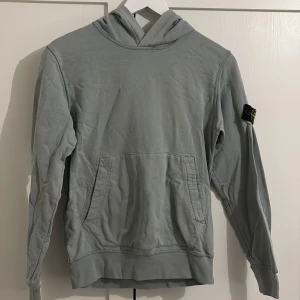 Grå hoodie från Stone Island - Säljer en stilren grå hoodie från Stone Island med deras ikoniska märke på ärmen. Tröjan har en klassisk känguruficka och en bekväm huva. Perfekt för en avslappnad stil. Tröjan är i bra skick och inköpt från NK kids i Stockholm