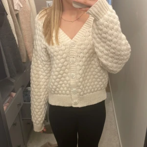 Vit stickad kofta - Mysig vit stickad kofta med knappar framtill. Koftan har en v-ringad design och långa ärmar. Den har ett fint strukturerat mönster.