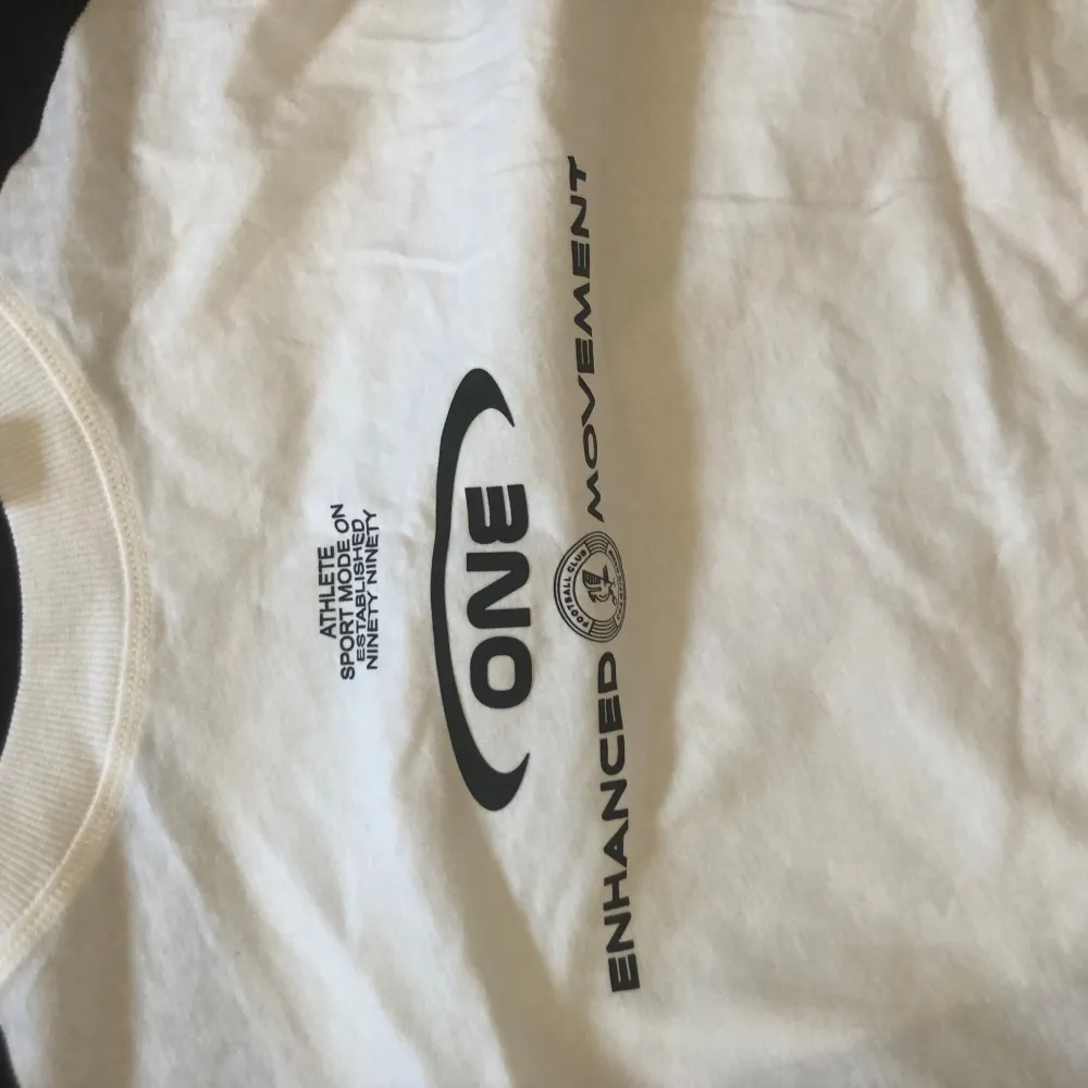 Säljer en vit och svart t-shirt med tryck 'ONE ENHANCED MOVEMENT' på framsidan. T-shirten har korta ärmar och en rund halsringning. Perfekt för en sportig look eller avslappnad stil.. T-paidat.