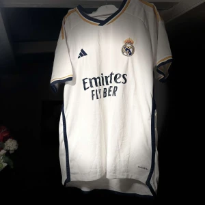 Vit Real Madrid fotbollströja - Säljer en vit Real Madrid fotbollströja från Adidas med korta ärmar. Tröjan har klubbens emblem på bröstet och sponsorlogga 'Emirates Fly Better' på framsidan. Den har blå och guld detaljer vid ärmslut och krage. Perfekt för alla Real Madrid-fans!