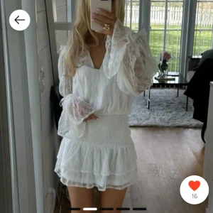 Vit spetsklänning med volanger - Klänningen har långa ärmar och en v-ringad design. Den smockade midjan ger en fin passform och kjolen har en volangkant. Perfekt för sommarens alla tillfällen! Säljer då jag valt en annan till studenten