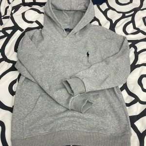 Grå hoodie från Polo Ralph Lauren - Säljer en stilren grå hoodie från Polo Ralph Lauren med det klassiska broderade logotypen på bröstet. Tröjan har en bekväm passform och är perfekt för en avslappnad look. Den har en praktisk känguruficka och ribbade muddar vid ärmslut och nederkant.