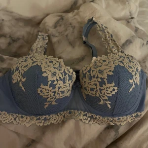 Blå bh med spetsdetaljer - Säljer en elegant blå bh med vackra spetsdetaljer i vit. Bh:n har justerbara axelband och en fin blommig spetsdesign som ger en romantisk känsla. Perfekt för att känna sig extra fin under kläderna. Bh:n är ifrån Intimissmi! Säljer för att den är för liten och därmed aldrig använd! 