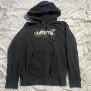 Svart hoodie från Levi's - Säljer en svart hoodie från Levi's med en camouflagemönstrad logga på bröstet. Tröjan har en klassisk känguruficka och justerbar huva med snören. Perfekt för en avslappnad stil.