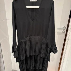 Svart från NA-KD - Snygg svart klänning från NA-KD med v-ringning och långa ärmar. Klänningen har plisserade detaljer vid midjan och ärmsluten som ger en elegant touch. Perfekt för en stilren look.