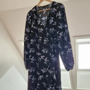Svart blommig omlottklänning - Snygg svart omlottklänning med blommigt mönster i vitt och blått. Klänningen har långa ärmar och en v-ringad design som ger en elegant look. Perfekt för både vardag och festliga tillfällen.