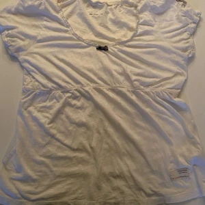 Vit kortärmad topp med rosett - Säljer en söt vit topp från Odd Molly med korta ärmar och en liten rosett framtill. Toppen har en fin spetsdetalj runt halsringningen och en lätt rynkad midja för en feminin touch.