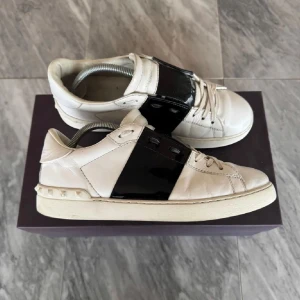 Valentino sneakers med svart detalj - Snygga vita sneakers från Valentino med en bred svart rem över snörningen. Skorna har en stilren design med nitar på hälen och är tillverkade i skinn. Perfekta för en trendig look.                              Det finns dock ingen box och inget kvitto men det fins bevis på att dom e äkta så hör av er vid minsta fundering.!