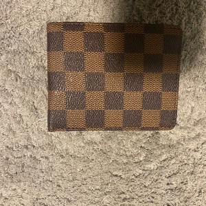 Rutig plånbok i brunt och beige - Snygg plånbok med klassiskt rutmönster i brunt och beige. Perfekt för att hålla ordning på kort och kontanter. Den har flera fack på insidan för enkel organisering. Priset är ej hugget i sten