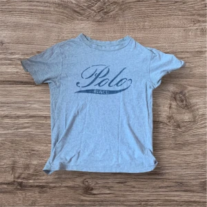 Grå t-shirt från Ralph Lauren - Säljer en grå t-shirt från Ralph Lauren med 'Polo' tryckt i blått på framsidan. T-shirten har en klassisk rund hals och korta ärmar. Perfekt för en avslappnad stil.