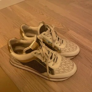 Beige och guld sneakers från Michael Kors - Snygga sneakers i beige med guldiga detaljer från Michael Kors. Skorna har ett mönster på ovansidan och snörning framtill.