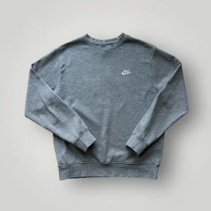 Grå sweatshirt från Nike - Nike Crewneck Sweatshirt – Storlek M  Snygg och stilren grå Nike-tröja i klassisk crewneck-modell. Tillverkad i mjukt och bekvämt material, perfekt för vardagsbruk eller avslappnade tillfällen. Tröjan har en diskret vit Nike-logga broderad på vänster bröst, vilket ger en minimalistisk och stilren look. Ribbstickade muddar vid ärmslut och nederkant ger en bra passform och extra komfort.  Detaljer: 	•	Märke: Nike 	•	Färg: Grå 	•	Storlek: M 	•	Passform: Normal 