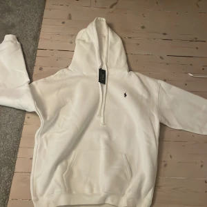 Vit hoodie från Ralph Lauren - Säljer en stilren vit hoodie från Ralph Lauren med en klassisk design. Hoodien har en liten broderad logga på bröstet och en praktisk känguruficka framtill. Perfekt för en avslappnad look. Helt ny. Passar M-L.
