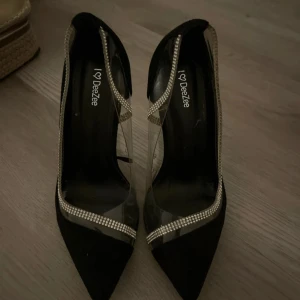 Svarta pumps från DeeZee med glittriga detaljer - Eleganta svarta pumps från DeeZee med spetsig tå och klack. Skorna har transparenta paneler och är dekorerade med glittriga detaljer längs kanterna, vilket ger en lyxig känsla.