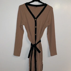 Beige ribbad klänning från Shein - Snygg beige ribbad klänning från Shein med svarta detaljer och knappar framtill. Klänningen har långa ärmar och ett knytband i midjan för en smickrande passform. Perfekt för en stilren look!