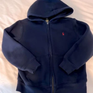 Mörkblå hoodie från Ralph Lauren - Säljer en snygg mörkblå hoodie från Ralph Lauren med dragkedja och det klassiska röda logotypen på bröstet. Perfekt för kyliga dagar med sin mjuka och bekväma passform. Huvan ger extra värme och stil. Passar både till vardags och avslappnade tillfällen.