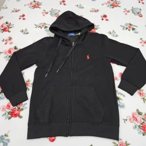 Svart hoodie från Polo Ralph Lauren - Säljer en stilren svart hoodie från Polo Ralph Lauren. Den har en dragkedja framtill och en klassisk huva med snörning. Tröjan är tillverkad i 100% bomull och har det ikoniska röda logotypbroderiet på bröstet. Perfekt för en avslappnad look.