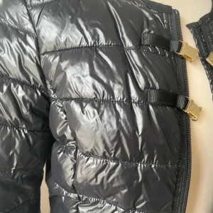 Svart dunjacka från Ilse Jacobsen - Snygg svart dunjacka från Ilse Jacobsen med dragkedja och stilrena spännen framtill. Jackan är vattenavvisande och fylld med konstgjort dun, perfekt för kyliga dagar. Den har en modern look med quiltad design och långa ärmar.