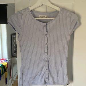 Jättesöt topp med knappar - Säljer en ljusblå ribbad topp från Abercrombie med knappar framtill. Toppen har korta ärmar och en rund halsringning. Perfekt för en avslappnad stil.