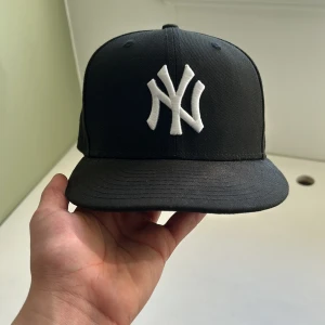 Svart fitted keps New Era - Snygg svart keps från New Era med vit broderad logotyp framtill. Kepsen har en klassisk design med böjd skärm och är i storlek 7 1/4 (57.7 cm). Perfekt för en stilren look.