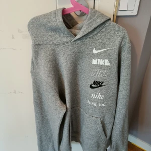 Grå hoodie från Nike - Säljer en grå hoodie från Nike med flera logotyper tryckta på framsidan. Tröjan har en klassisk design med huva och långa ärmar. Perfekt för en avslappnad stil. den är aldrig använd då jag fick den i present och inte riktigt var min stil