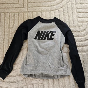 Grå och svart tröja från Nike - Säljer en grå och svart tröja från Nike med långärmade svarta ärmar och stort Nike-tryck på framsidan. Fickor sitter vid sidan. Perfekt för en sportig look!
