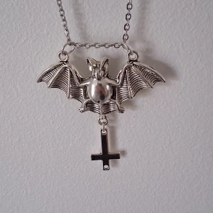 Halsband med fladdermus och kors - Unikt halsband i silverfärg med en detaljrik fladdermus och ett hängande kors. Kedjan är tunn och smidig, vilket gör smycket lätt att bära. Perfekt för att ge en edgy touch till din stil.