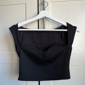 Svart ribbad topp - Snygg svart topp med ribbad textur och en stilren design. Toppen har en croppad passform och breda axelband som ger en modern look. Perfekt för att styla med höga jeans eller kjol.