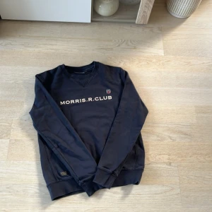 Mörkblå tröja från Morris R. Club - Säljer en snygg mörkblå tröja från Morris R. Club med text på bröstet och ett litet emblem. Tröjan har långa ärmar och ribbade muddar vid ärmslut och nederkant. Perfekt för en stilren look!