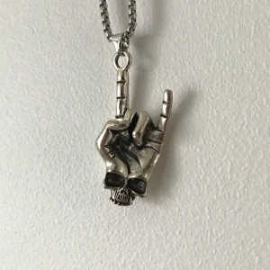 Halsband med handformad berlock - Coolt halsband med en berlock i form av en hand som gör rocktecknet. Berlocken är i silverfärg och har en dödskalle vid basen. Kedjan är robust och matchar berlockens stil. Perfekt för att ge en edgy touch till din outfit.