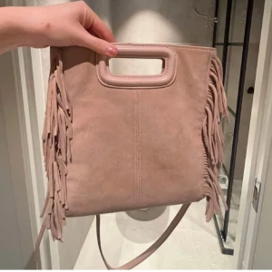 Rosa axelväska med fransar - Snygg rosa axelväska i mocka med coola fransdetaljer på sidorna. Väskan har ett praktiskt handtag och en avtagbar axelrem för olika bärstilar. Perfekt för att ge din outfit en bohemisk touch!