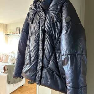 Blå  pufferjacka med huva - Snygg blå pufferjacka med huva och dragkedja. Jackan har en glansig yta och stilrena detaljer med knappar vid kragen. Perfekt för kyliga dagar när du vill hålla dig varm och trendig.