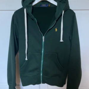 Mörkgrön hoodie från Polo Ralph Lauren - Snygg mörkgrön hoodie från Polo Ralph Lauren med gul broderad logga på bröstet. Tröjan har huva med vita,helt ny och skriv frågor vid funderingar!!!!