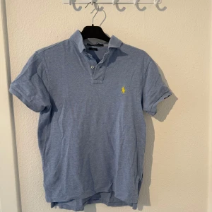 Blå pikétröja från Polo Ralph Lauren - Snygg blå pikétröja från Polo Ralph Lauren i custom fit. Klassisk krage, korta ärmar och ikoniska gula loggan broderad på bröstet. 