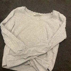 Ljusgrå långärmad topp med spets H&M - Supersöt ljusgrå långärmad topp från H&M med spetsdetaljer runt halsringningen och en liten rosett framtill. Mjuk och skön bomullskänsla, perfekt för dig som gillar en enkel men ändå söt stil.