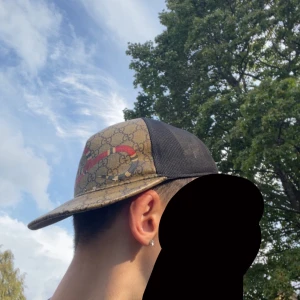 Gucci keps med GG-mönster och orm - Säljer en Gucci keps med klassiskt GG-monogram i beige och brunt, prydd med en röd och svart orm framtill. Baksidan är i svart mesh och justerbar med grön-röd randig rem. Perfekt för dig som vill sticka ut med en ikonisk accessoar. Tillverkas ej längre.