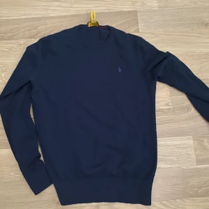 Marinblå tröja från Polo Ralph Lauren - Snygg marinblå tröja från Polo Ralph Lauren med diskret broderad logga på bröstet. Tröjan har rund hals och långa ärmar, perfekt för dig som gillar klassisk stil. Materialet känns mjukt och skönt mot huden.
