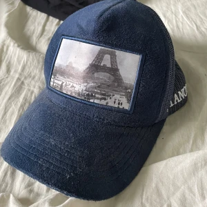 Blå Pianotti keps med Eiffeltornet - Snygg blå keps från Pianotti med mesh på sidorna och broderad logga. Framtill finns en stor patch med ett svartvitt foto av Eiffeltornet. Tyvärr är en av plumparna på spännet bort och några lätt skadad