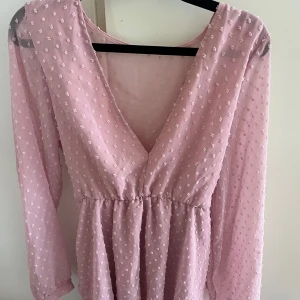 Ljusrosa prickig kortklänning med bälte - Säljer en söt ljusrosa kortklänning med prickigt mönster och transparenta långärmar. Klänningen har v-ringad front, markerad midja och ett matchande bälte. Materialet är lätt och luftigt, perfekt för vår och sommar. Superfin till sneakers eller sandaler!