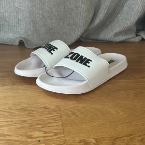 Vita Zone sandaler med tryck helt nya - Säljer ett par fräscha vita sandaler från Zone med svart text på ovandelen. De har en bred rem över foten och en platt sula med mönstrad undersida för extra grepp. Perfekta att glida runt i under varma dagar. Materialet är syntet och designen är enkel men stilren.